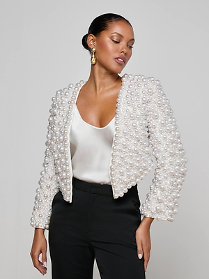 A woman wears the Belle Oversized Pearl Jacket by L'AGENCE—a statement piece with hand-sewn faux pearls—styled with a white satin top, black pants, gold earrings, and a sleek low bun against a plain background.