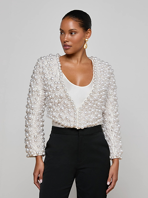 A woman models the L'AGENCE Belle Oversized Pearl Jacket, featuring hand-sewn faux pearls over a white top and high-waisted black pants. With sleek hair and gold earrings, she creates a striking look as she gazes off to the side.