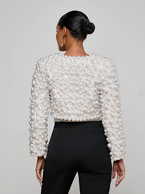 A woman with her hair in a neat bun and gold hoop earrings faces away, showcasing the L'AGENCE Belle Oversized Pearl Jacket—a white, long-sleeve statement piece with hand-sewn faux pearls—paired with high-waisted black pants.