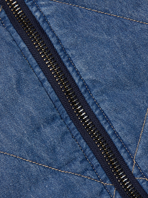 Close-up of the Ryah Bomber Jacket by L'AGENCE, featuring quilted stitching with visible detail and a black zipper running diagonally across blue lightweight denim fabric.