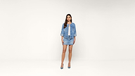 A woman stands against a plain white background, wearing a light blue denim jacket, matching denim mini skirt, white top, and blue high heels. She has long brown hair and a neutral expression.