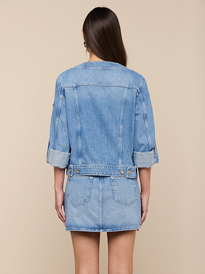 A woman with long brown hair, seen from behind, wears the Anouk Jean Jacket with rolled-up sleeves and a matching denim skirt with back pockets, standing against a beige background.