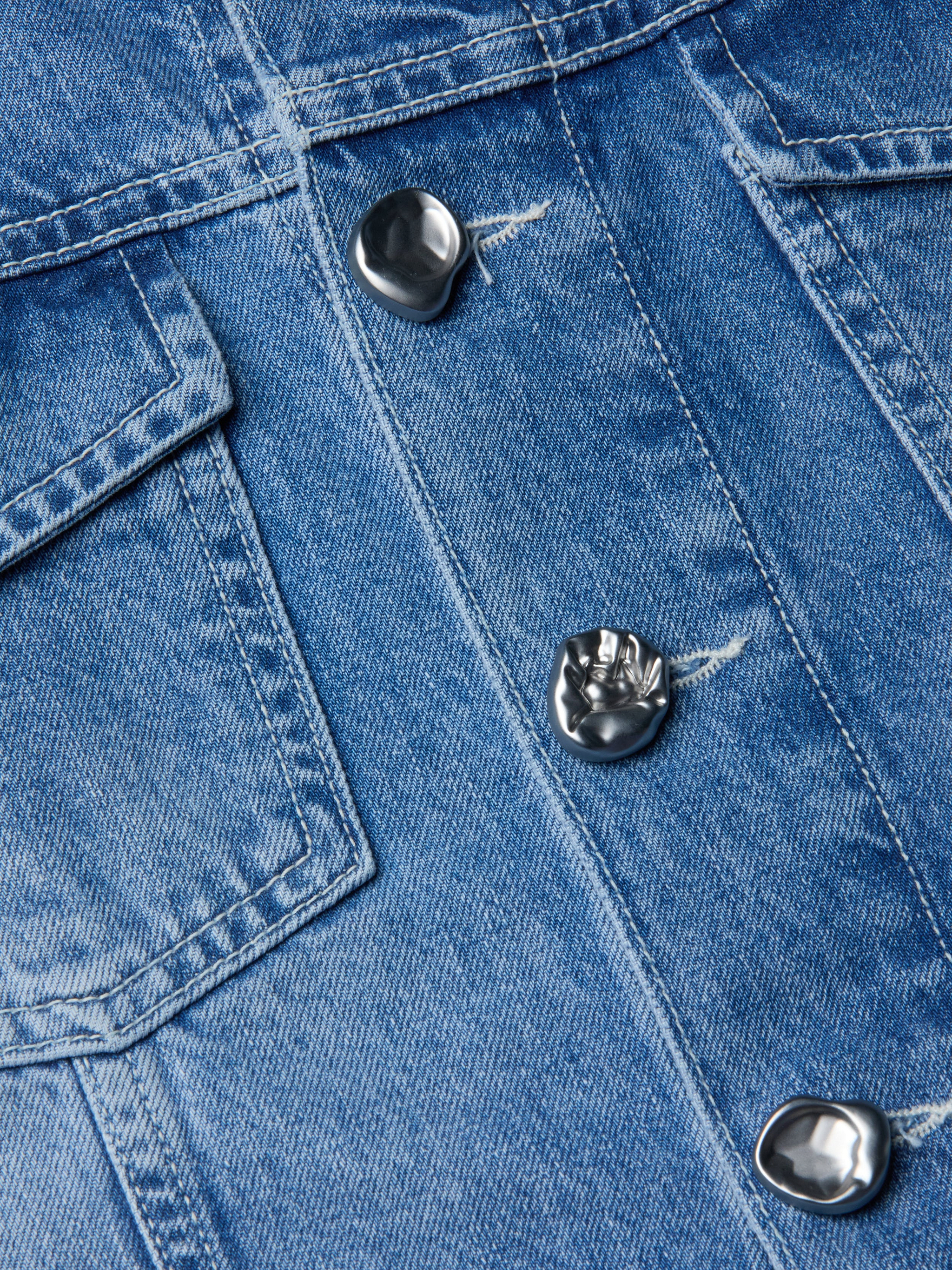 لقطة مقرّبة لجاكيت Anouk Jean Jacket المصنوع من قماش الدنيم النيلي مع أزرار معدنية فضية وغطاء جيب مرئي، حيث تظهر خامة الثوب المزخرفة والخياطة المفصلة.