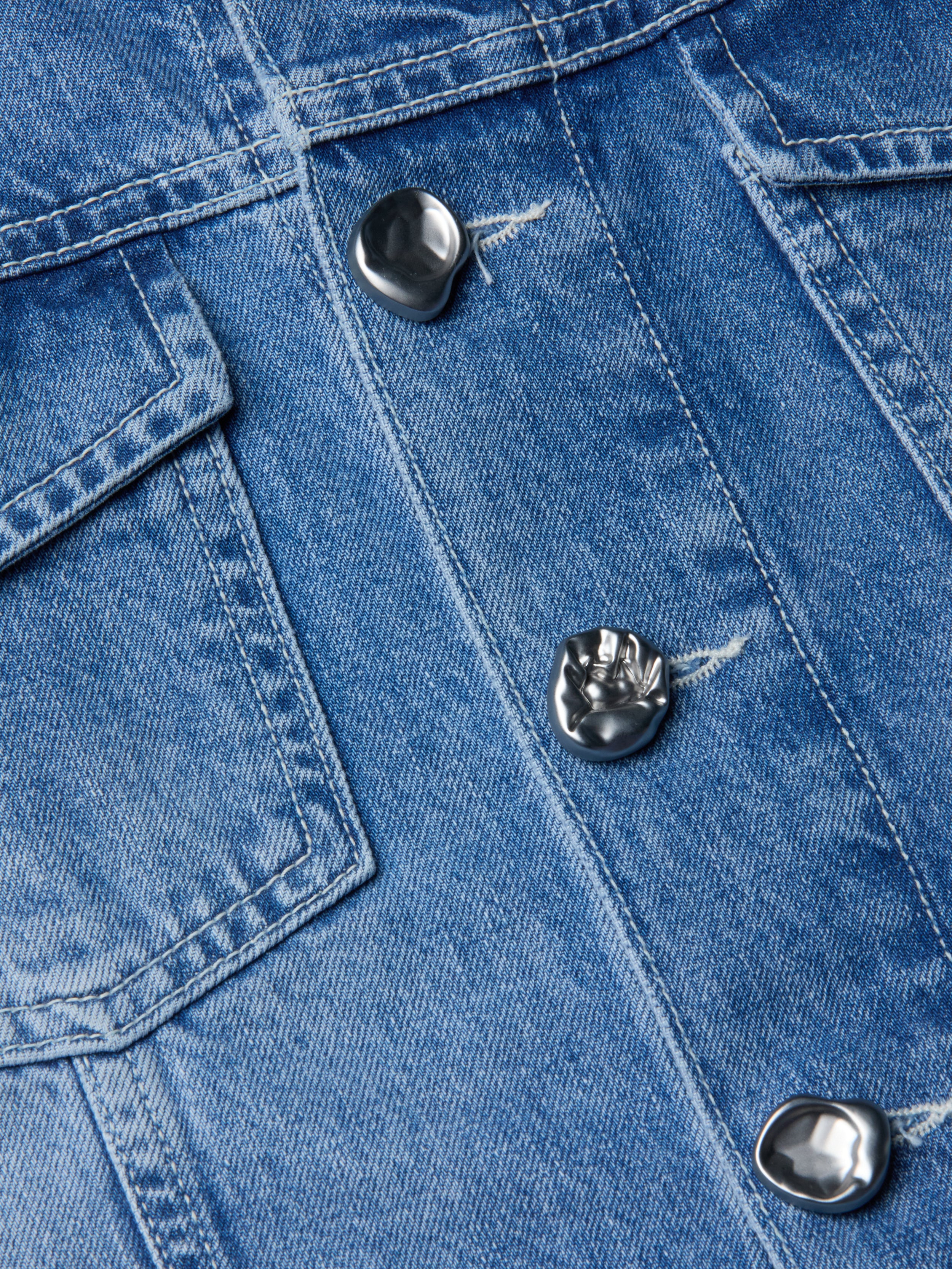 لقطة مقرّبة لجاكيت Anouk Jean Jacket المصنوع من قماش الدنيم النيلي مع أزرار معدنية فضية وغطاء جيب مرئي، حيث تظهر خامة الثوب المزخرفة والخياطة المفصلة.
