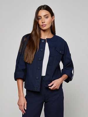 A woman with long brown hair models the Anouk Jean Jacket—a relaxed fit, collarless denim jacket in navy blue—over a white top and coordinating navy pants, standing against a plain light gray background.