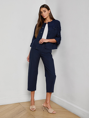 A woman with long brown hair stands indoors by a white wall, wearing the Anouk Jean Jacket in navy blue and matching high-waisted, wide-leg pants over a white top, styled with beige heeled sandals.