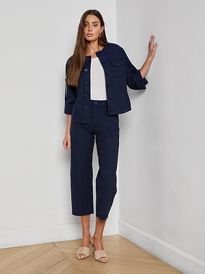 A woman with long brown hair stands indoors, leaning against a white wall. She wears the Anouk Jean Jacket in navy blue with a relaxed fit, matching navy cropped pants, a white top, and beige heeled sandals.