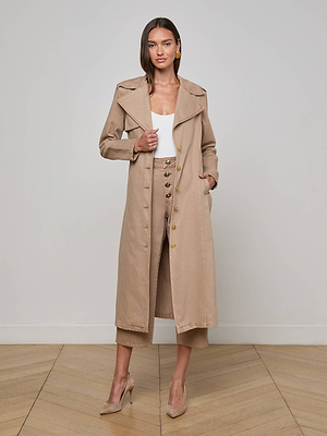 A woman stands indoors on a wood floor wearing the L'AGENCE Zavia Jean Trench Coat over a white top and tan high-waisted pants with large buttons, paired with tan heels. She poses with one hand in her pocket.