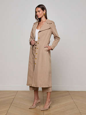 A woman with long brown hair poses indoors against a plain background, wearing L'AGENCE's Zavia Jean Trench Coat with an oversized collar and waist tie over a white top and beige pants, paired with beige high heels.