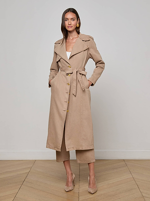 A woman stands indoors wearing the L'AGENCE Zavia Jean Trench Coat, featuring an oversized collar, waist tie belt, wide lapels, gold buttons, and pockets. She styles it with neutral heels and beige cropped pants against a plain background.