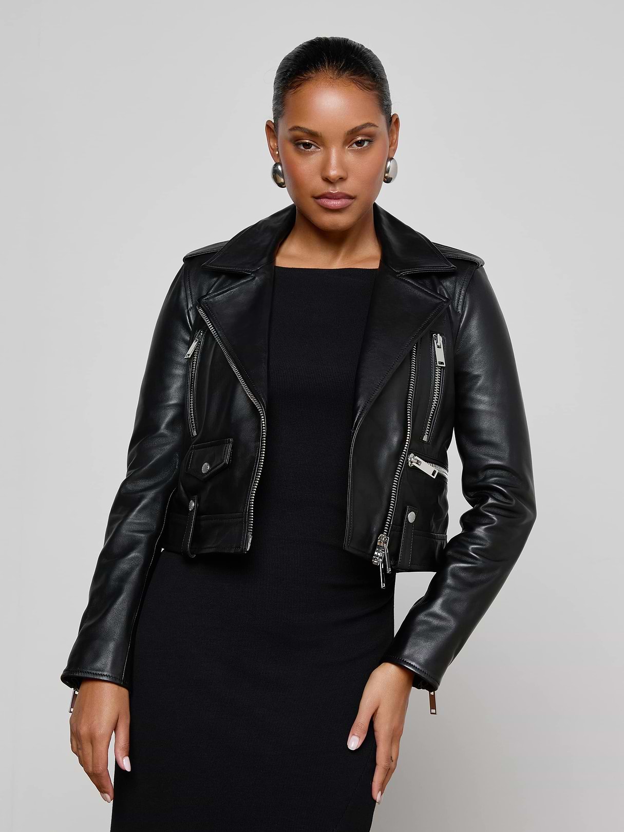 Wearing the Blakely Leather Biker Jacket over a black dress, a woman stands against a plain light background, looking confidently at the camera with one hand on her hip.