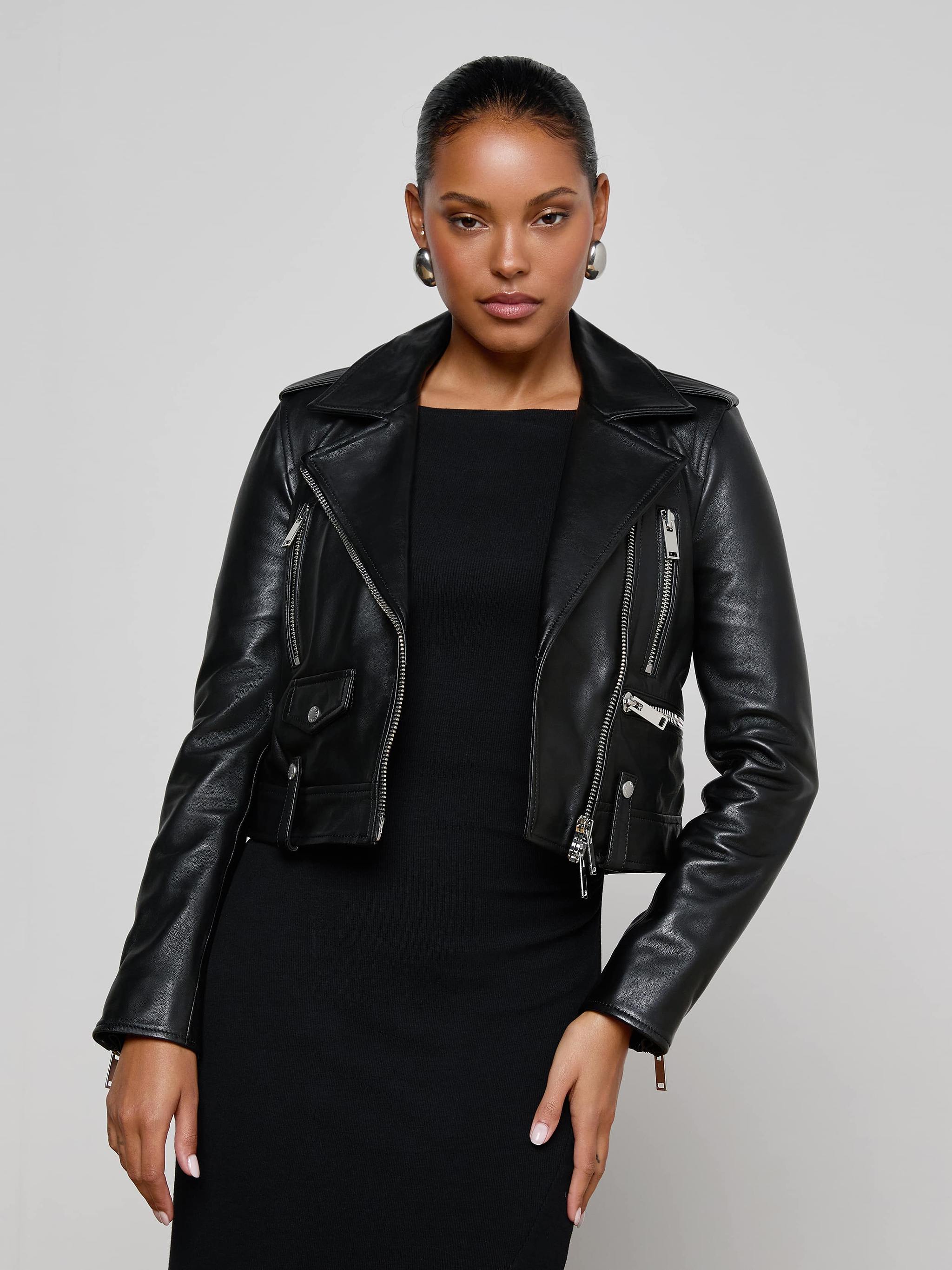 Wearing the Blakely Leather Biker Jacket over a black dress, a woman stands against a plain light background, looking confidently at the camera with one hand on her hip.