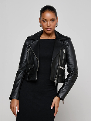A woman stands confidently against a plain gray background, wearing the Blakely Leather Biker Jacket over a black dress, her hand on her hip as she looks at the camera.