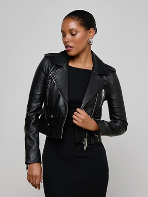 A woman with dark hair pulled back poses against a plain light background, wearing the Blakely Leather Biker Jacket—cropped black lambskin—over a fitted black dress, looking down as she adjusts the jacket’s zipper.