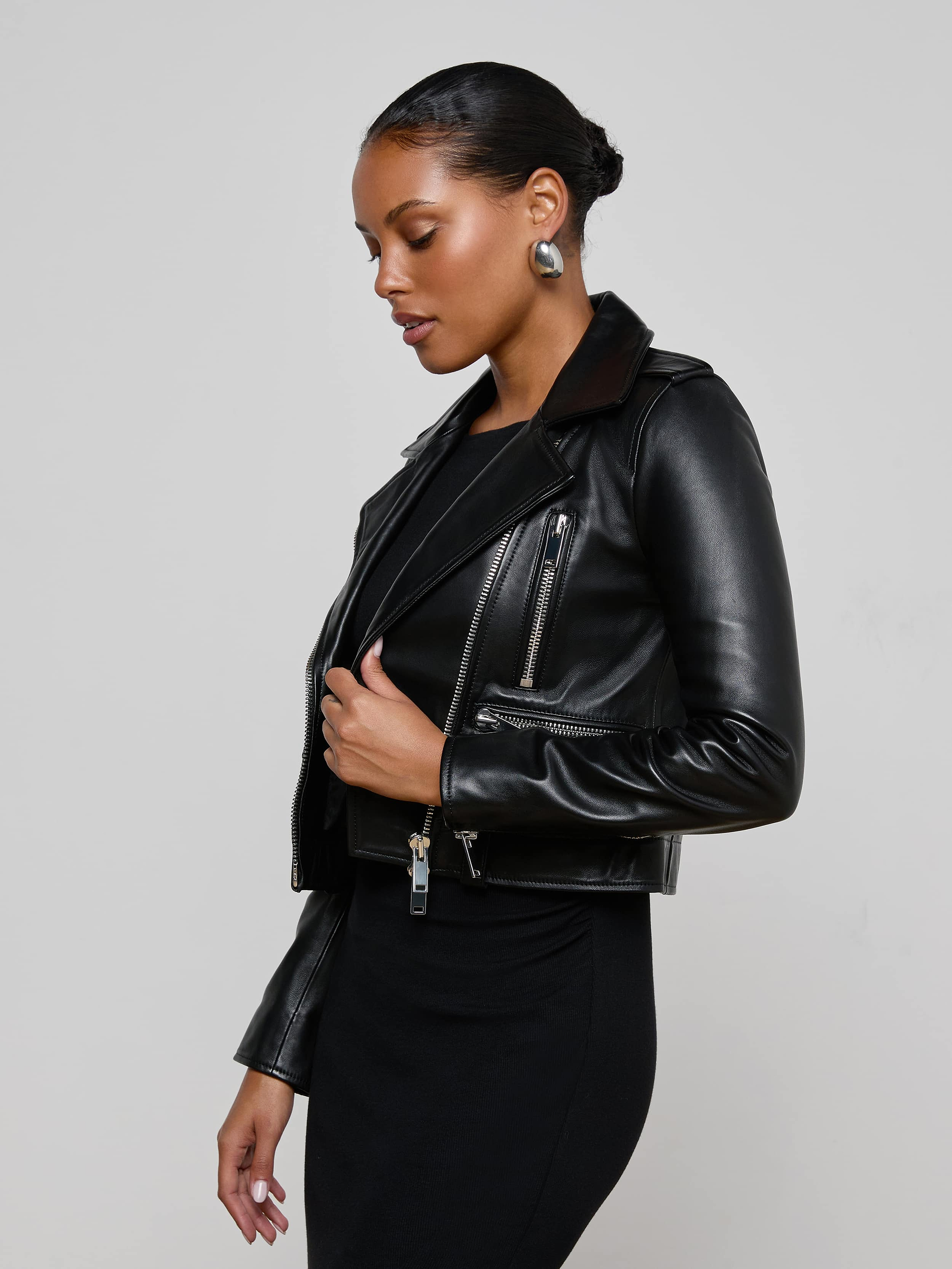 Wearing the Blakely Leather Biker Jacket over a black dress, a woman with short, styled hair and large round earrings stands against a plain light background. She gazes downward, her hand resting on the jacket’s lapel.