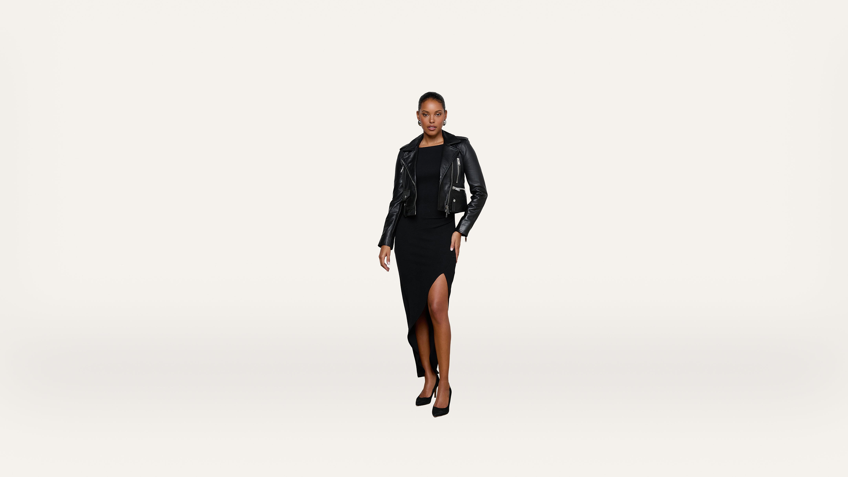 Woman in a black dress with a thigh-high slit, black leather jacket, and black heels stands confidently against a plain white background.