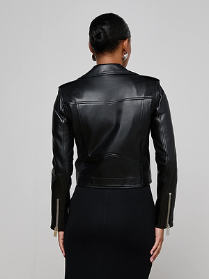 A woman faces away, wearing the Blakely Leather Biker Jacket—fitted black lambskin with visible seams and zippered cuffs—paired with a black skirt. Her hair is in a neat bun, and she accessorizes with stud earrings.