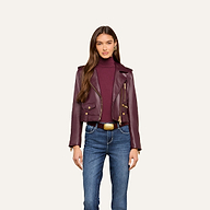 A woman stands facing forward against a plain, light background, wearing the Blakely Leather Biker Jacket in maroon lambskin over a matching turtleneck and blue jeans.