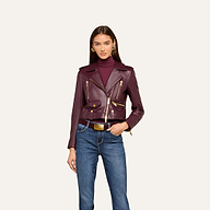 A woman wears the Blakely Leather Biker Jacket with a cropped fit over a burgundy turtleneck and blue jeans. She has long straight hair and gold earrings, standing against a plain light background.