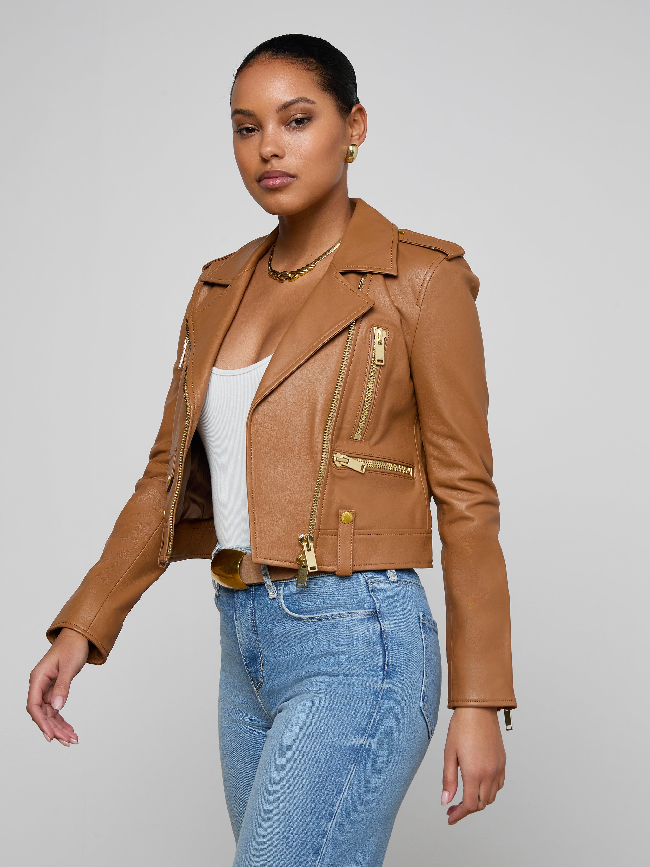 A woman stands confidently against a plain light background, wearing the Blakely Leather Biker Jacket in tan lambskin over a white top and blue jeans, looking directly at the camera.