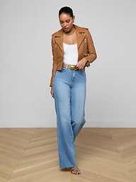A woman wearing the Blakely Leather Biker Jacket in brown, a white top, blue high-waisted wide-leg jeans, and sandals stands on a plain white background, looking down.