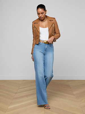 A woman stands indoors on a light wood floor against a plain white wall, wearing the Blakely Leather Biker Jacket in brown with a white top, light blue wide-leg jeans, beige belt, and heeled sandals.