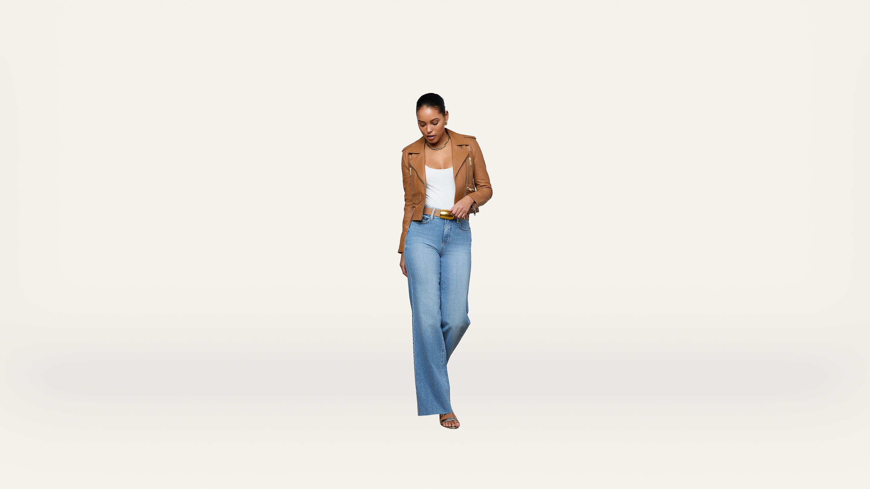 A woman stands against a plain white background, wearing a tan jacket, white top, light blue high-waisted jeans, and sandals. She is looking down, smiling, and has her hands in her pockets.