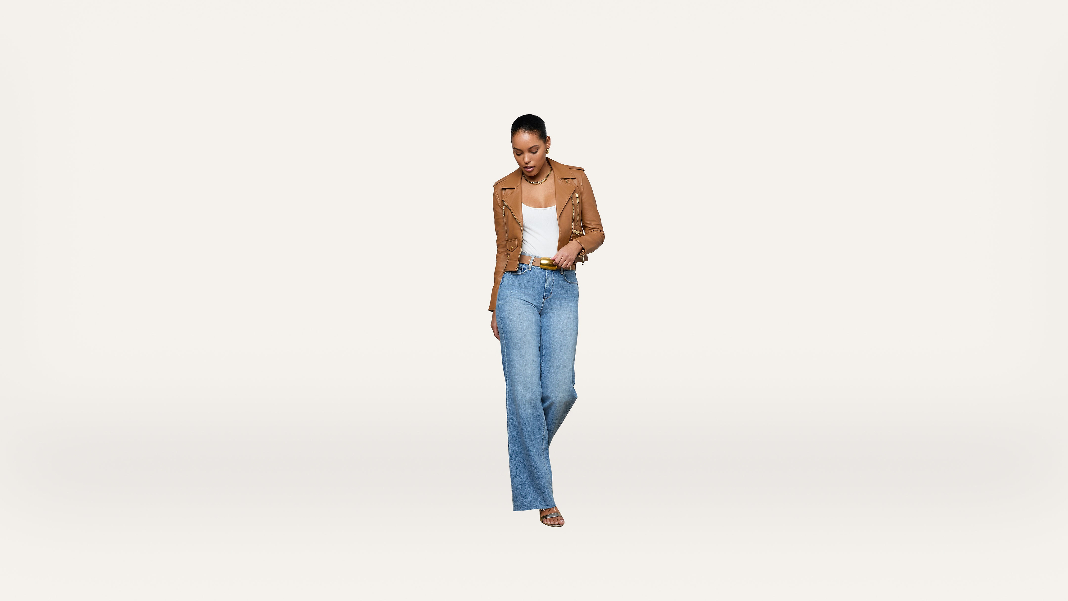 A woman stands against a plain white background, wearing a tan jacket, white top, light blue high-waisted jeans, and sandals. She is looking down, smiling, and has her hands in her pockets.