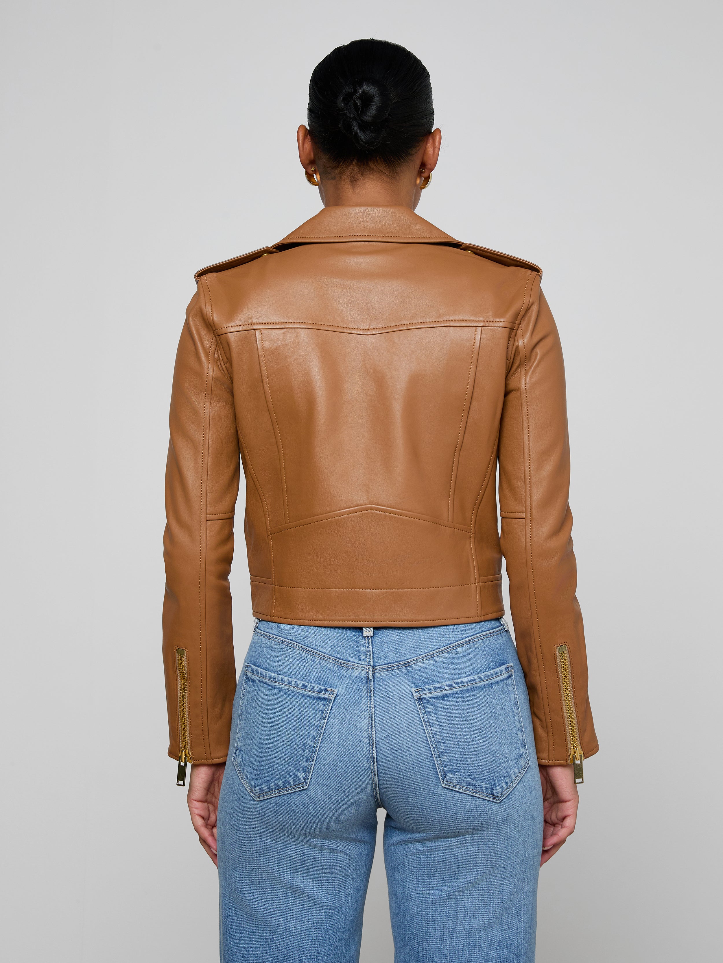 A person stands facing away, wearing the Blakely Leather Biker Jacket in brown lambskin and blue jeans. The background is plain and light-colored.