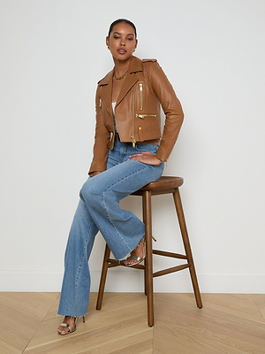 A woman with short hair and a confident look sits on a wooden stool against a white wall, wearing the Blakely Leather Biker Jacket, light blue flared jeans, gold hoop earrings, and strappy heels.