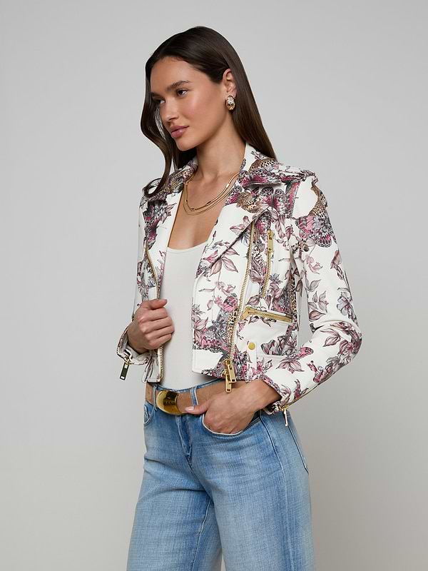 A woman wears the Blakely Leather Biker Jacket, featuring a white base and butterfly jungle print, layered over a white top with light blue jeans and accented by gold jewelry, all set against a plain light background.