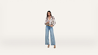 A woman stands against a plain white background, wearing wide-leg blue jeans, a floral blouse, beige heels, and a brown belt. She has straight hair and is looking at the camera with a slight smile.