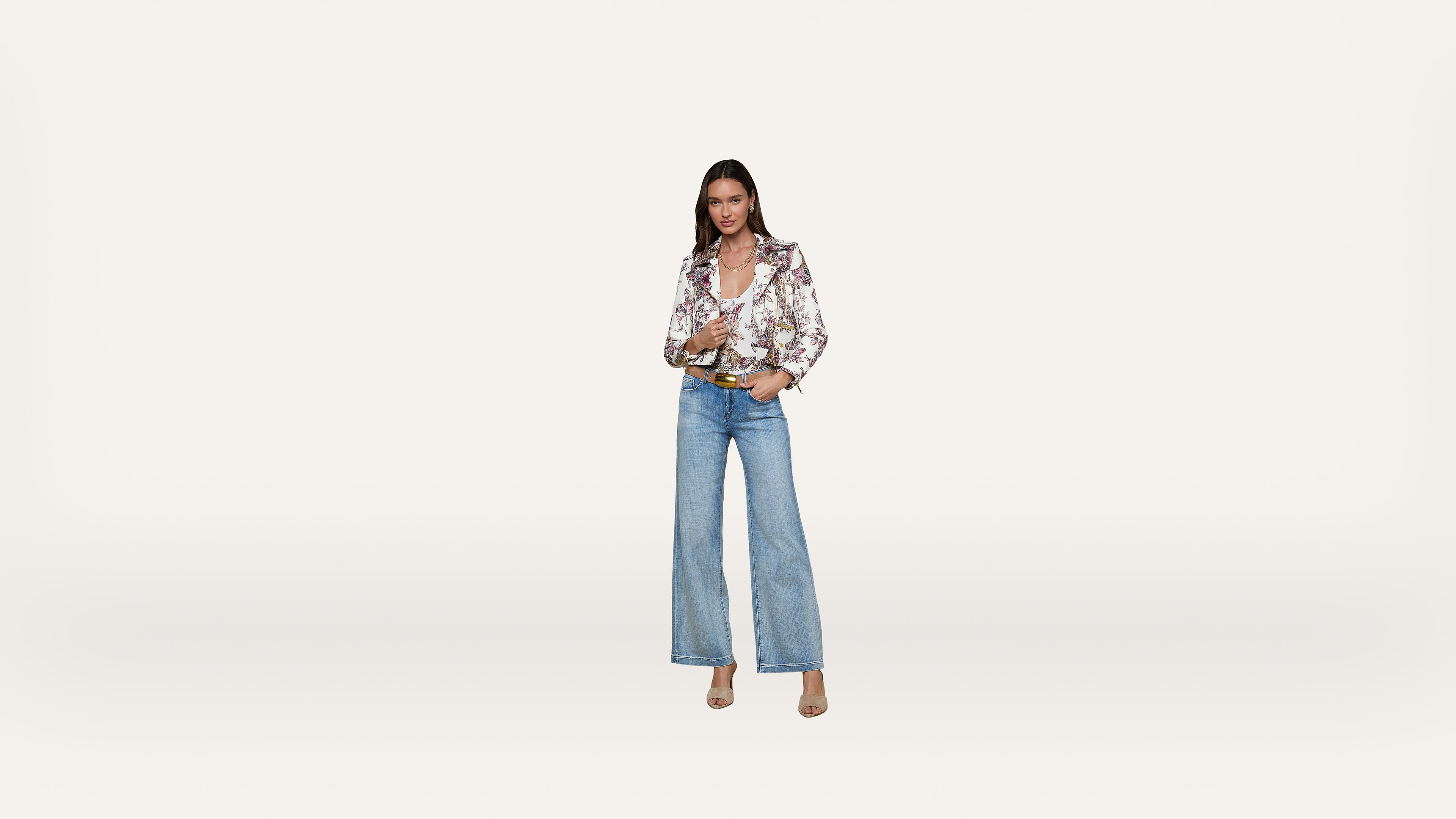 A woman stands against a plain white background, wearing wide-leg blue jeans, a floral blouse, beige heels, and a brown belt. She has straight hair and is looking at the camera with a slight smile.