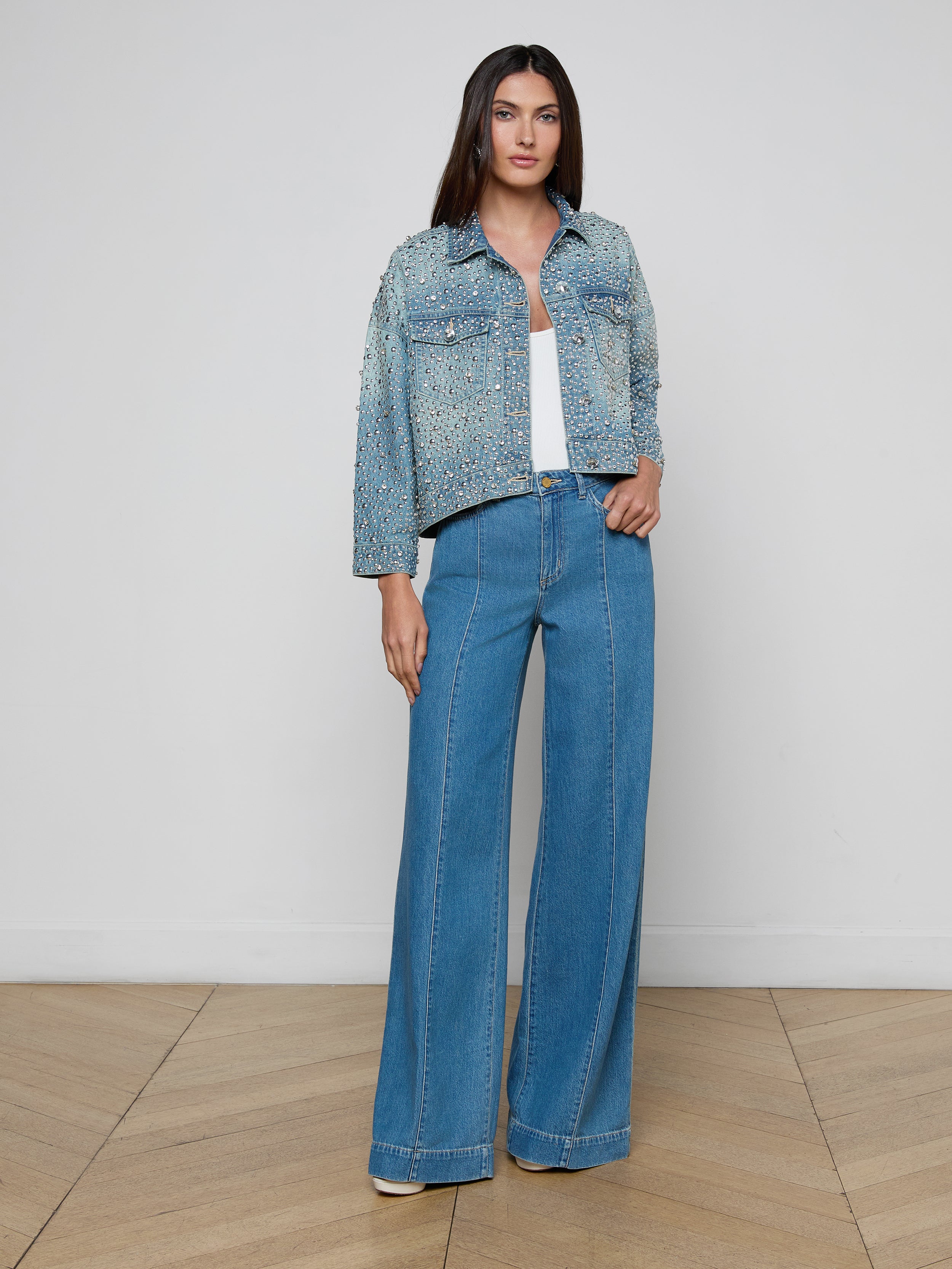 A woman with long straight hair poses indoors against a white wall, wearing the Vangie Embellished Jean Jacket over a white top and blue wide-leg jeans on a wooden floor.