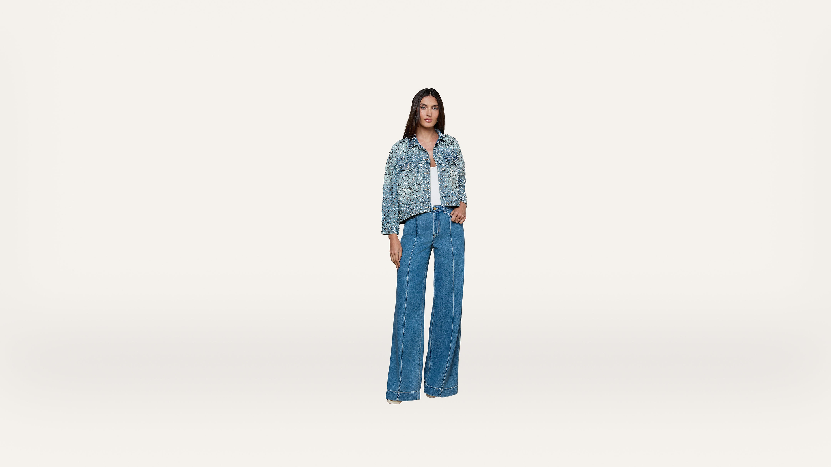 A woman stands against a plain white background, wearing a blue denim jacket over a white top and wide-leg blue jeans. She is facing forward with her arms relaxed by her sides.