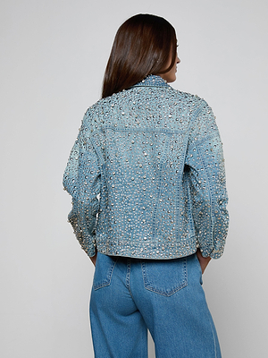 Seen from behind, a person with long brown hair wears the Vangie Embellished Jean Jacket featuring crystal details and a cropped high-low hem, paired with blue jeans against a plain gray background.