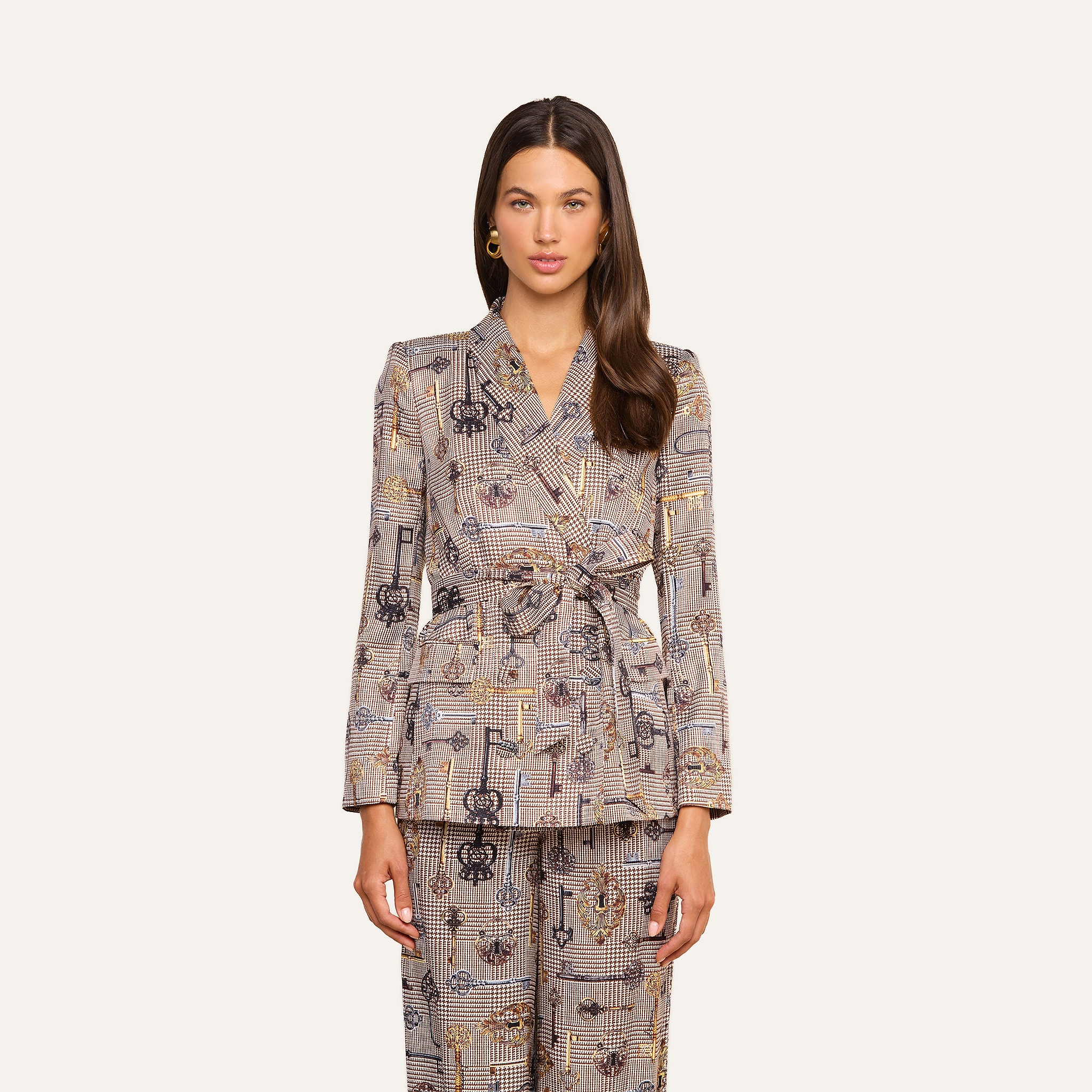A woman stands facing forward wearing the Aven Wrap Blazer—a modern wrap-front blazer paired with matching patterned pants and a belted waist. She has long brown hair and gold earrings, set against a neutral background.