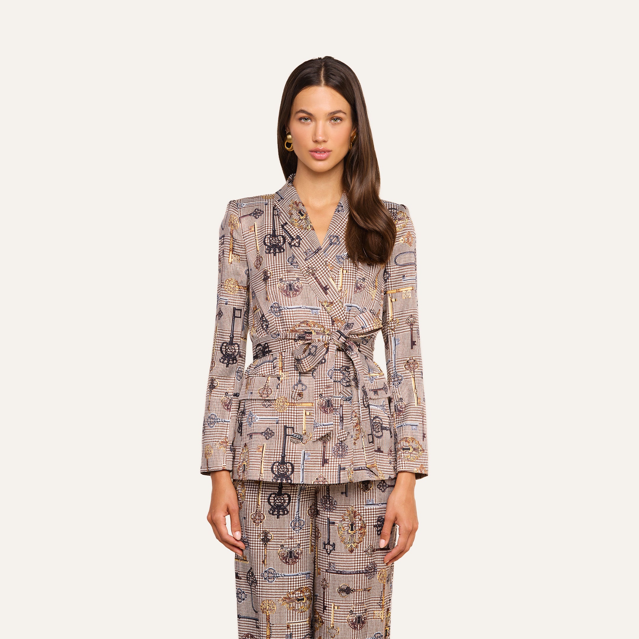 A woman stands facing forward wearing the Aven Wrap Blazer—a modern wrap-front blazer paired with matching patterned pants and a belted waist. She has long brown hair and gold earrings, set against a neutral background.