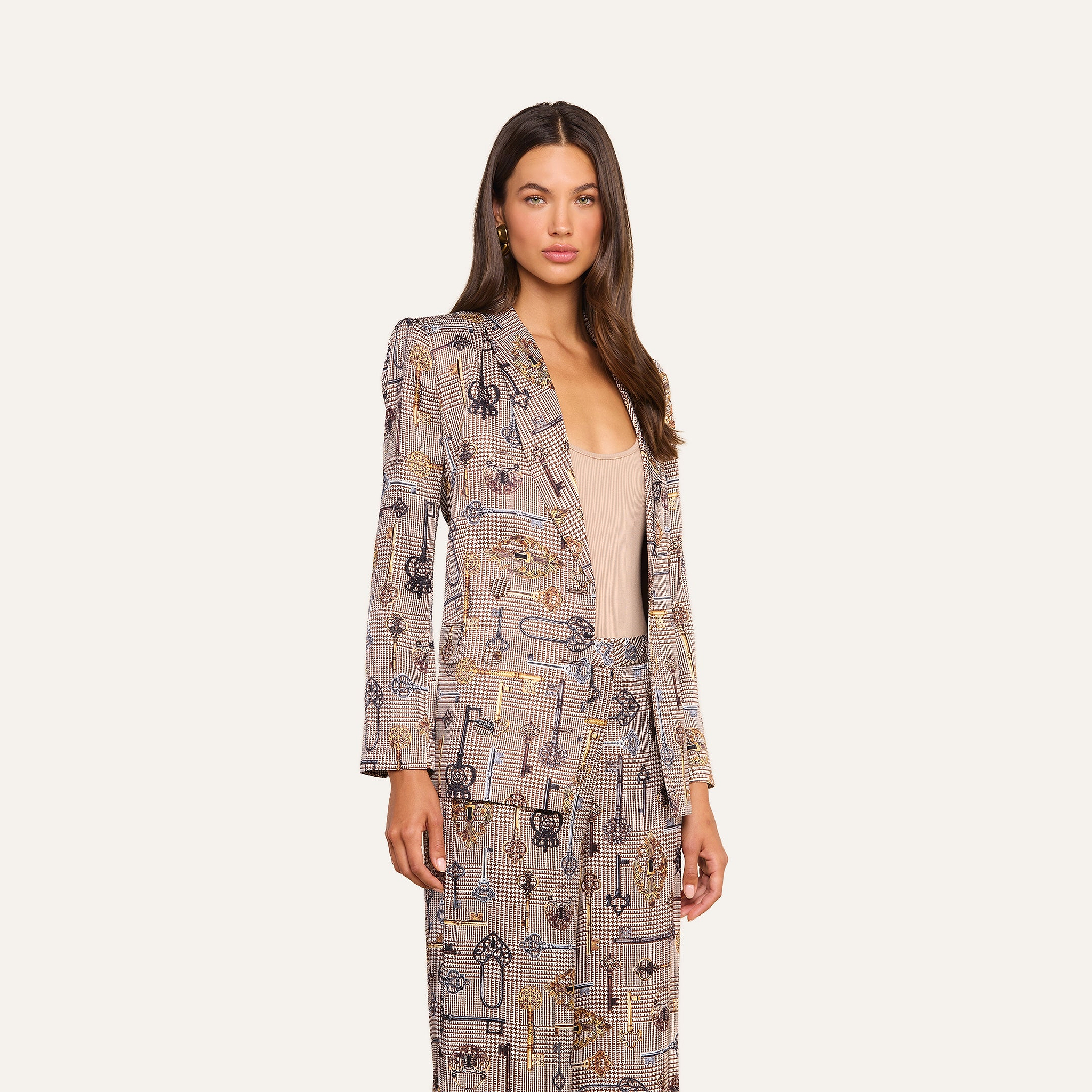 A woman stands facing forward in the Aven Wrap Blazer, a brown houndstooth patterned suit, layered over a light beige top. With one hand at her side and an off-white background, the modern suiting look is accentuated.
