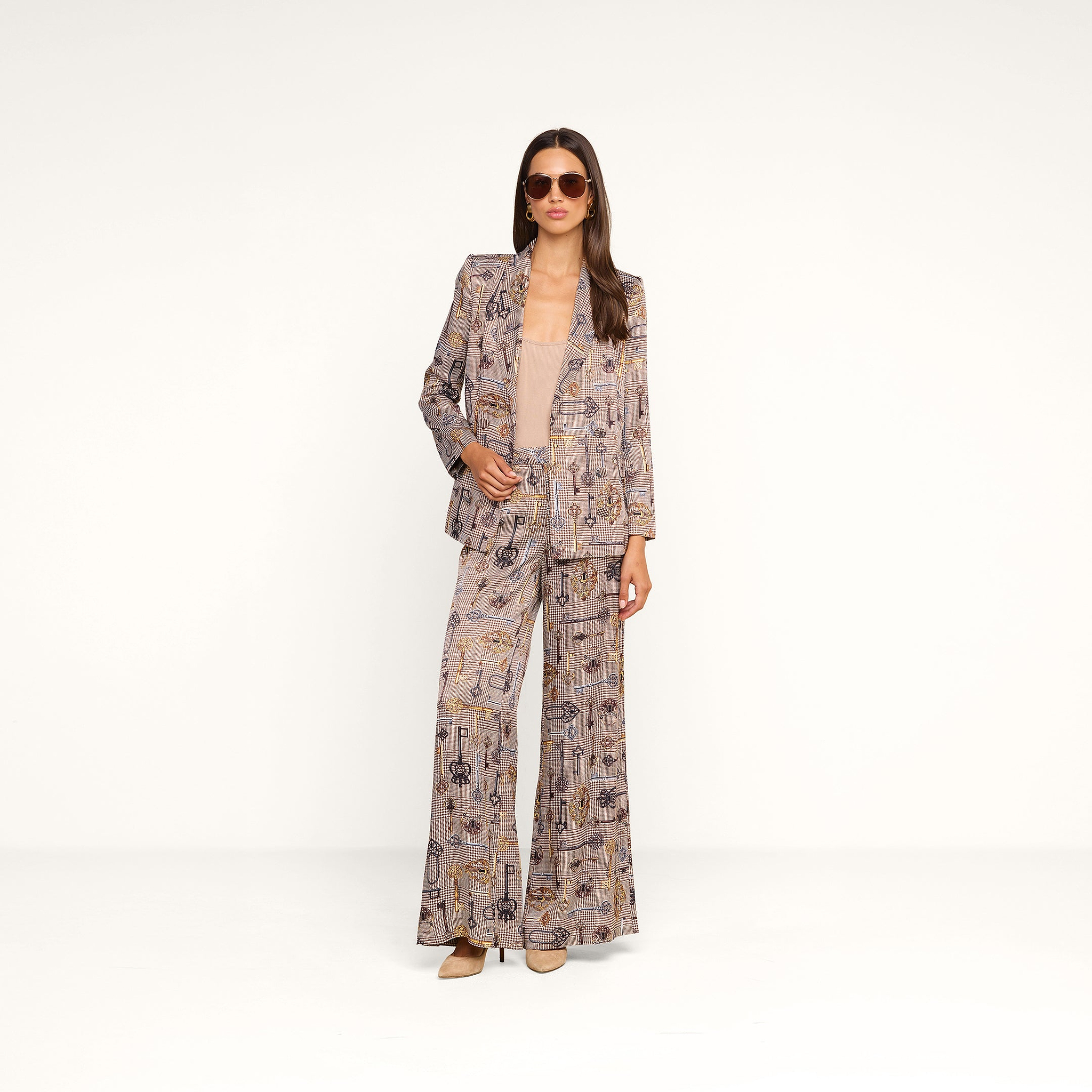 A woman stands confidently against a plain white background, wearing the Aven Wrap Blazer in a brown houndstooth pattern, paired with matching wide-leg pants, a tan top, heels, and sunglasses.