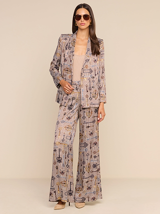 A woman stands confidently with one hand on her hip, dressed in the Aven Wrap Blazer— a modern brown houndstooth wrap-front blazer—styled with a beige top and coordinating wide-leg pants. She accessorizes with sunglasses, hoop earrings, and tan shoes, posing against a plain beige background.