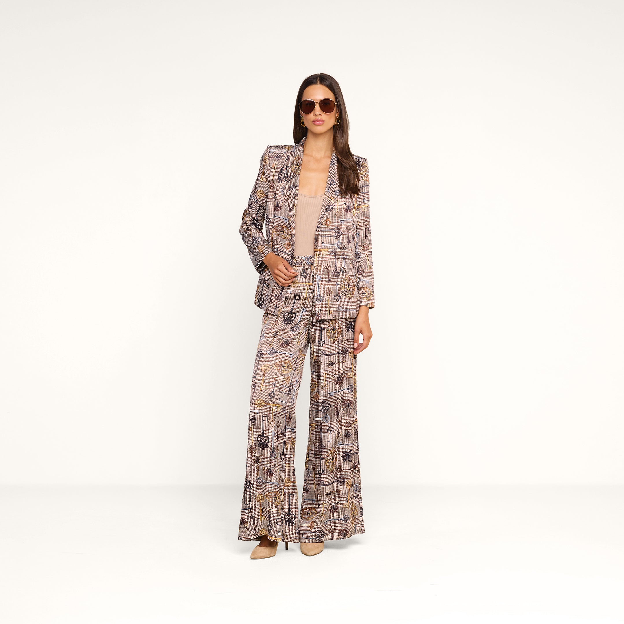 A woman stands confidently against a plain white background, wearing the Aven Wrap Blazer in a brown houndstooth pattern, paired with matching wide-leg pants, a tan top, heels, and sunglasses.