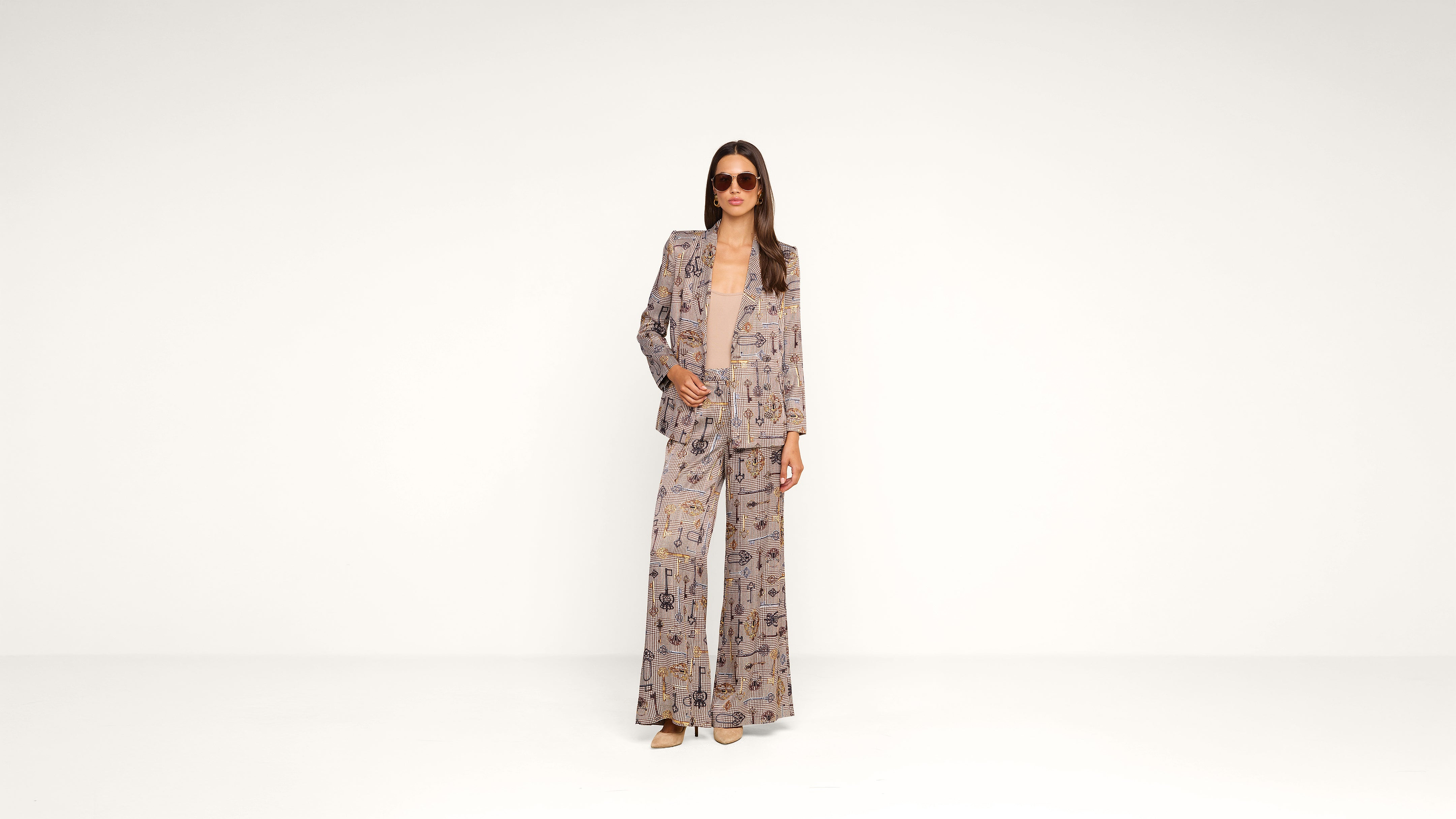 Woman wearing sunglasses, a patterned suit jacket, and wide-leg trousers stands against a plain white background. She has straight brown hair and is posing confidently with one hand in her pocket.