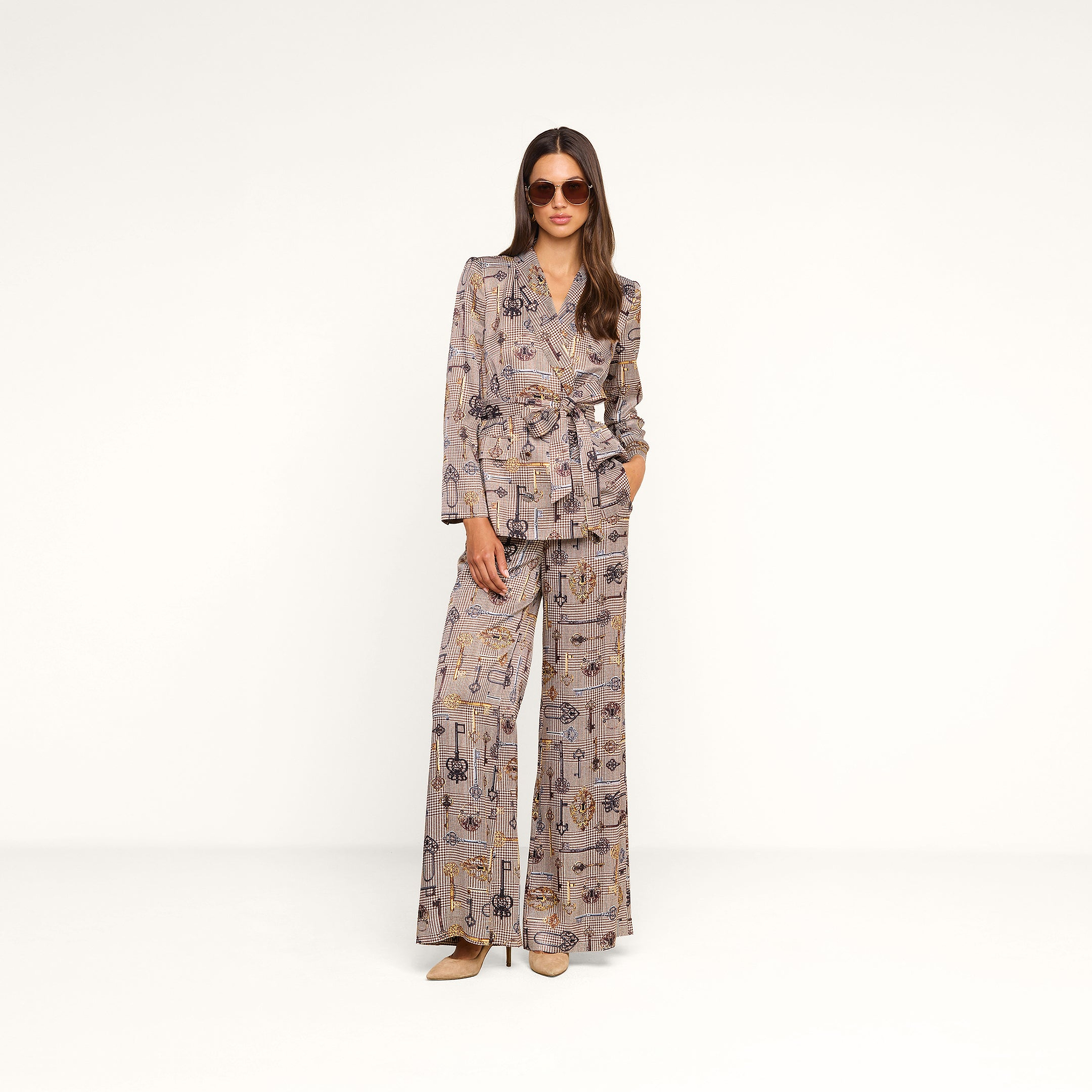 Dressed in the Aven Wrap Blazer and matching wide-leg houndstooth trousers, this woman accessorizes with sunglasses as she stands against a plain white background, creating a modern suiting look.