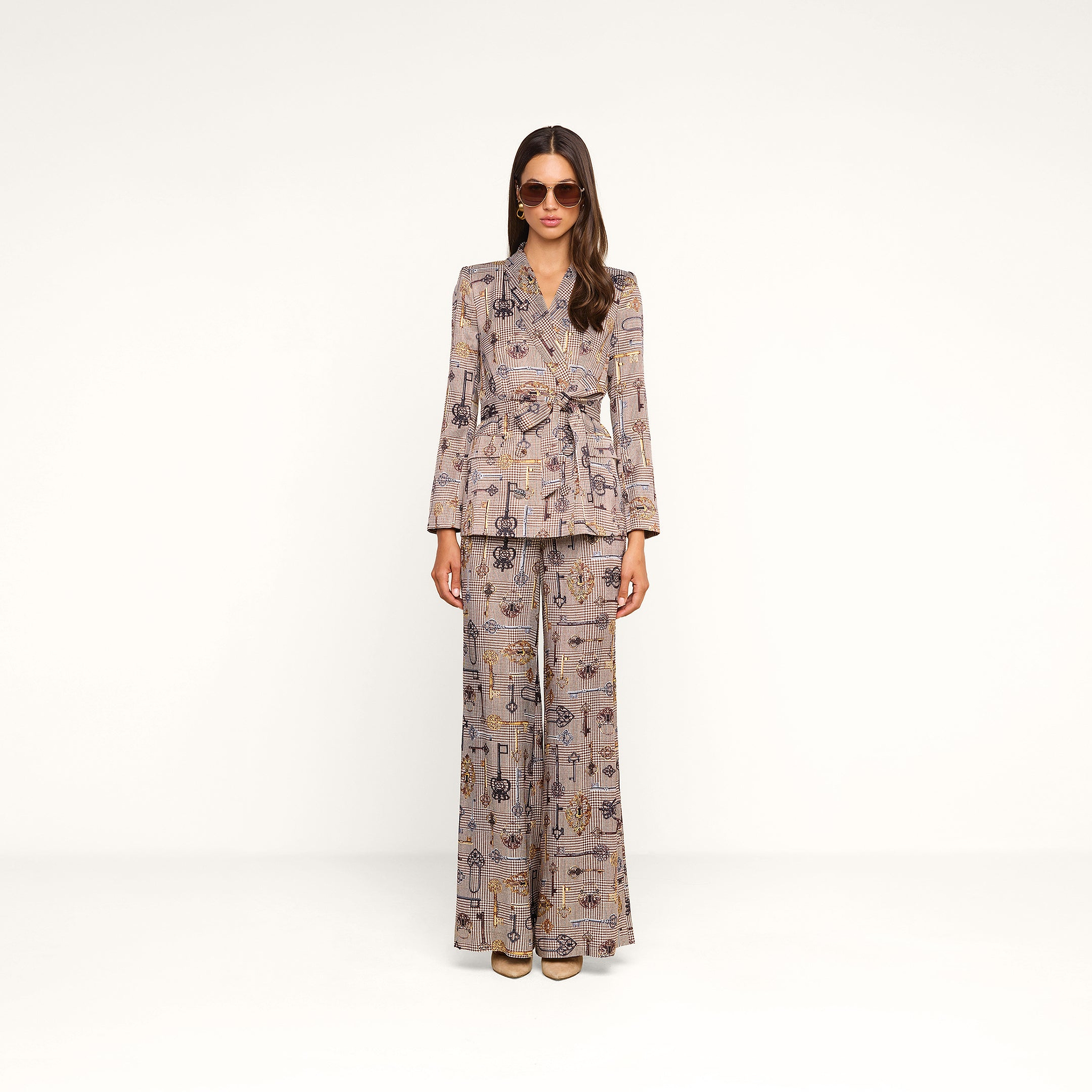 A woman wearing sunglasses and a patterned brown houndstooth pantsuit with the Aven Wrap Blazer stands against a plain white background, facing the camera with her arms at her sides.