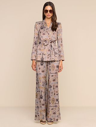 A woman wearing the Aven Wrap Blazer and matching wide-leg pants in a brown houndstooth pattern, paired with sunglasses and posed against a plain beige background—a sophisticated take on modern suiting.