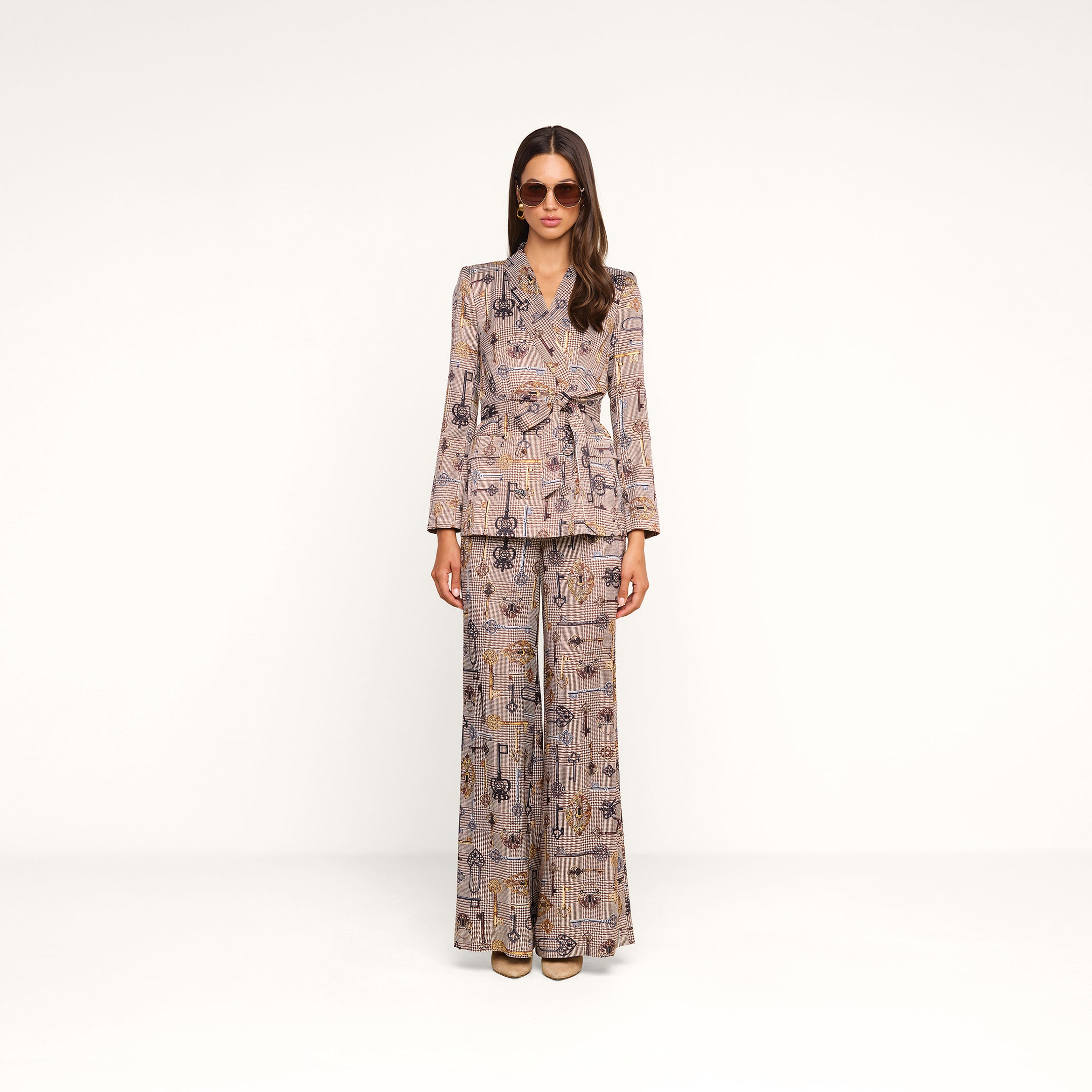 A woman wearing sunglasses and a patterned brown houndstooth pantsuit with the Aven Wrap Blazer stands against a plain white background, facing the camera with her arms at her sides.
