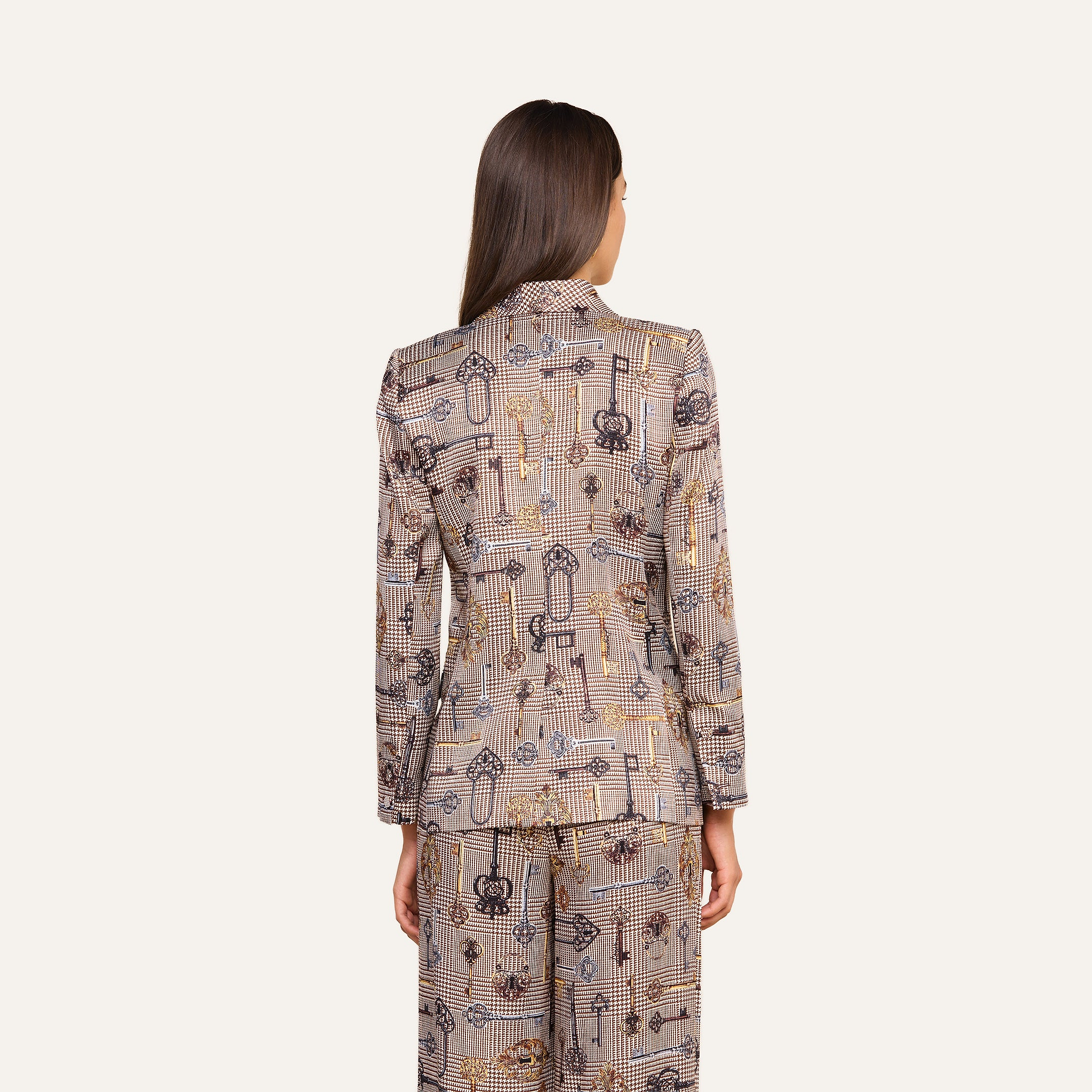 A person with long brown hair, seen from behind, wears the Aven Wrap Blazer and matching pants, both featuring abstract geometric and floral designs. The background is plain light beige.