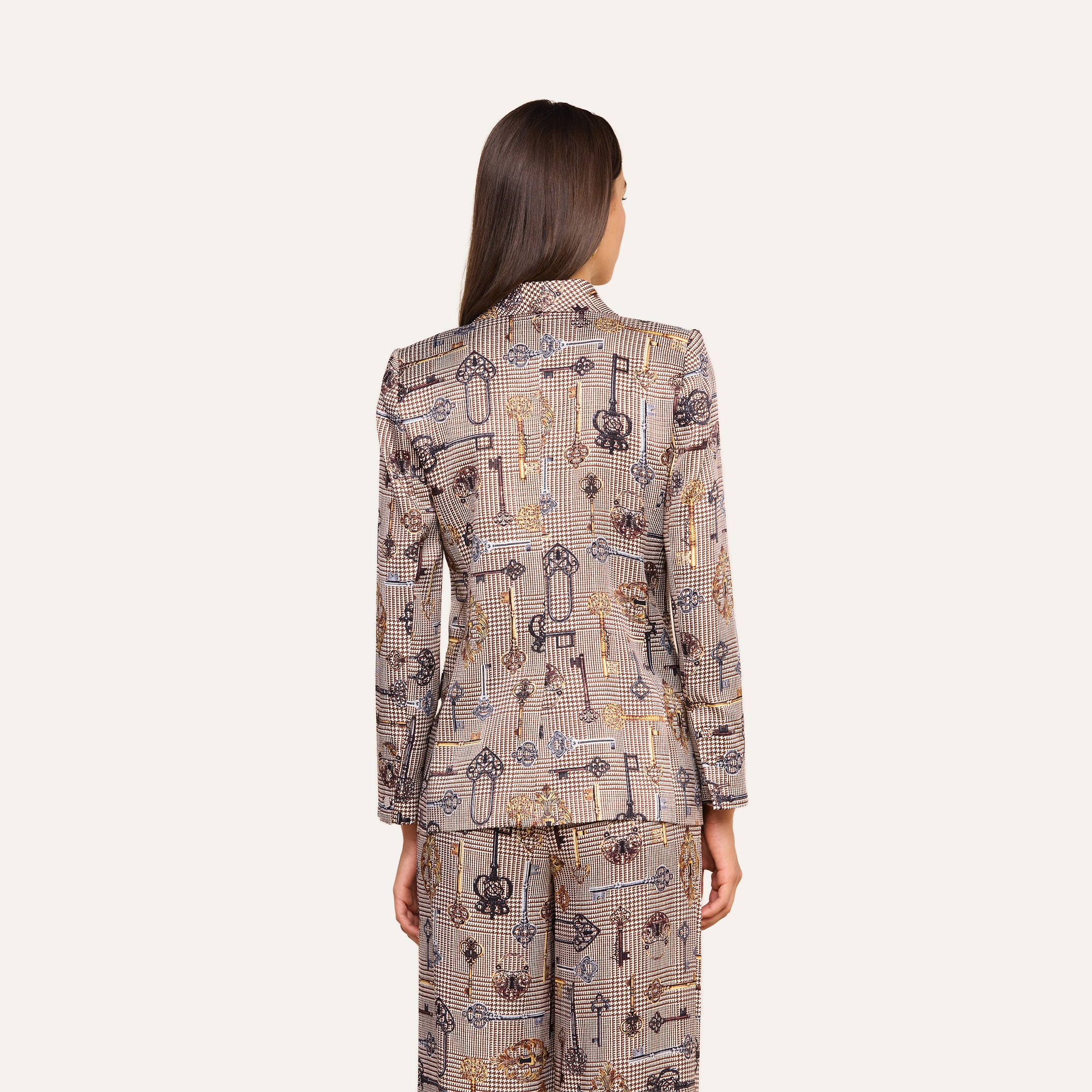 A person with long brown hair, seen from behind, wears the Aven Wrap Blazer and matching pants, both featuring abstract geometric and floral designs. The background is plain light beige.