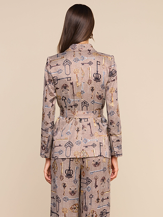 A woman stands with her back to the camera, wearing the Aven Wrap Blazer—a modern, belted jacket featuring a patterned wrap-front design—paired with matching pants adorned with abstract key and lock motifs on a neutral background.